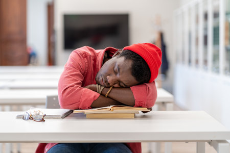 Lazy African male student falling asleep during study in library, sitting at desk sleeping on booksの写真素材