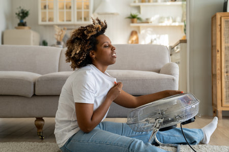 Overheated woman sitting on floor in living room using electric fan during extreme summer heatの写真素材