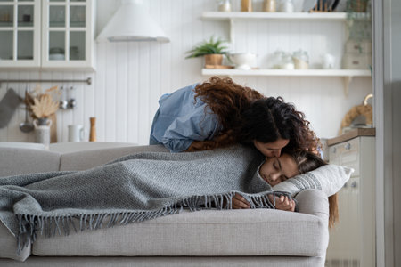 Young loving mother kissing daughter sleeping peacefully on couch during day nap time at homeの写真素材