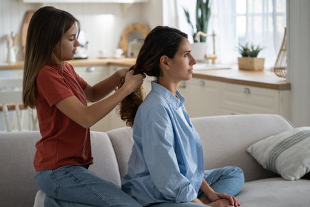 Daughter braiding mothers long wavy hair, mom and child enjoying time with each other at home