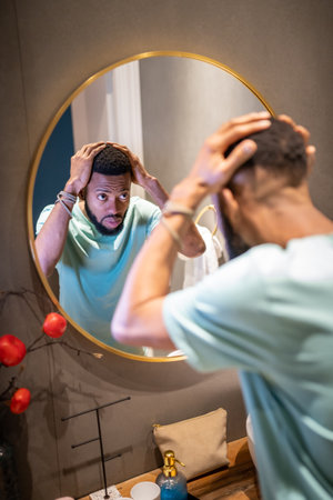 Upset young African American man looking in bathroom mirror touching hair worrying about baldingの写真素材
