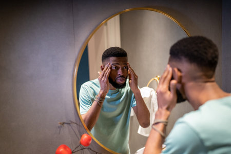 Worried african american man looks at mirror, touches facial skin anxious about wrinkles.
