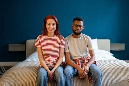 Portrait of happy married interracial diverse couple smiling at camera while sitting together on bedの写真素材
