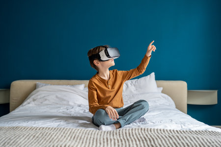 Child boy wearing virtual reality headset sitting on bed at home exploring metaverse. VR conceptの写真素材