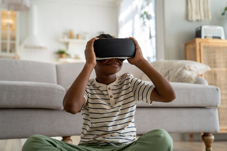 Obsessed child black boy experiencing 3D virtual reality in VR goggles sits at home Gadget addictionの写真素材