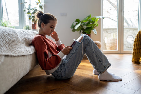 Upset teen girl sitting on floor near bed using smartphone at home, teenagers and gadget addictionの写真素材