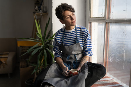 Optimistic Italian woman artist or craftsman with tea cup sits on windowsill and looks out window
