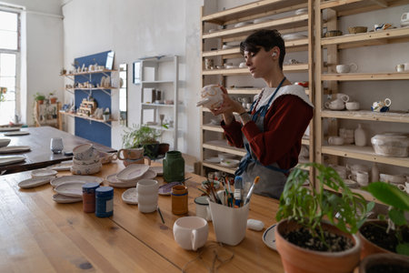 Concentrated woman potter holding paintbrush, painting on clay mug in workshopの写真素材
