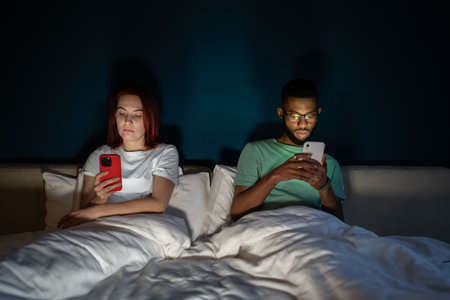 Young multiracial couple lying in bed using smartphones not talkingの写真素材
