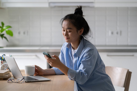 Smiling Asian girl freelancer holding smartphone chatting with friend while working from homeの写真素材