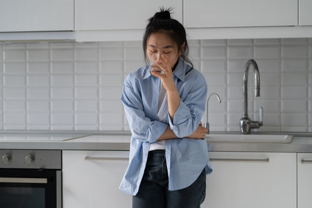 Stressed young Asian woman standing in kitchen drinking water calming down after panic attackの写真素材