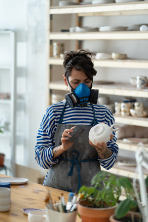 Woman ceramist wearing respirator polishing pottery in workshop, female potter at workplace