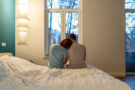 Family couple man and woman sitting together in bed hugging looking at window in bedroom at home.の写真素材