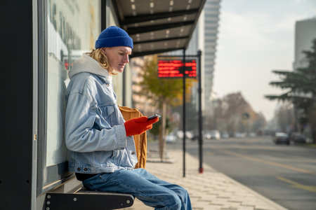 Caucasian millennial guy student with smartphone at bus station in early morning,の写真素材