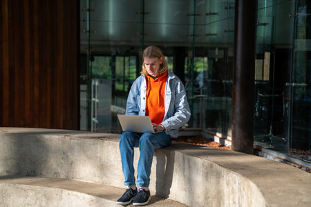 Student guy sitting with laptop against University building, studying online outdoorsの写真素材