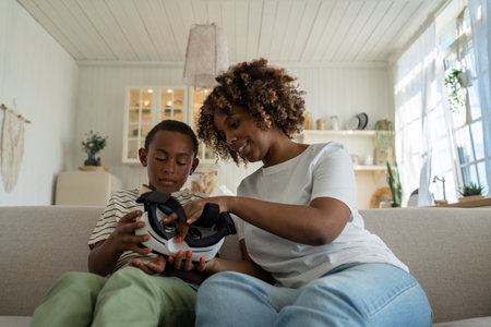 African american mom shows explains to son how use virtual reality glasses sits on couch at home.