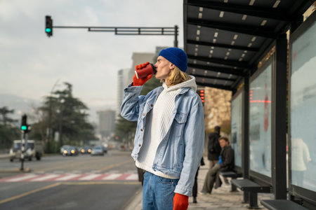 Stylish guy waiting bus standing on bus stop in downtown and drinking coffee.
