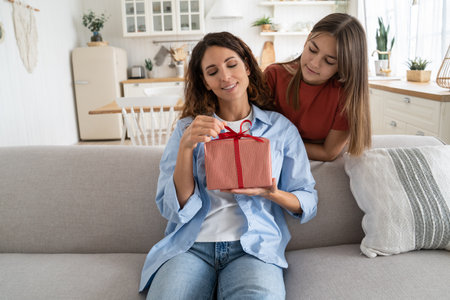 Excited young woman mother getting wrapped gift box present from daughter while sitting on sofaの写真素材