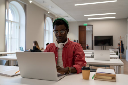 Puzzled student black hipster man sits at table with textbooks laptop studying in lecture classroom.の写真素材