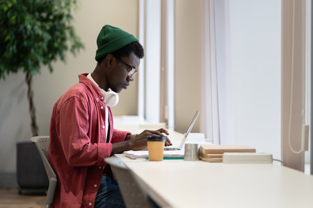 Focused concentrated black freelancer hipster guy sits at desk typing laptop keyboard in coworking.の写真素材