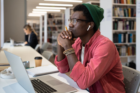 Thoughtful black male student wearing wireless earbuds sitting at desk with laptop in libraryの写真素材