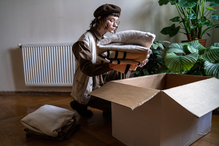 Caring volunteer girl in eyeglasses puts used clothes in cardboard box. Conscious consumption thingsの写真素材
