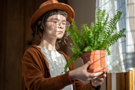 Hipster girl plant lover standing by window at home holding potted Boston fern houseplantの写真素材