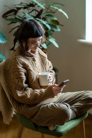 Sad teen girl holding cup of tea using phone feeling anxious while scrolling on social mediaの写真素材