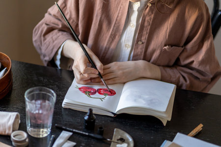 Female artist sitting at table holding paintbrush painting in sketchbook