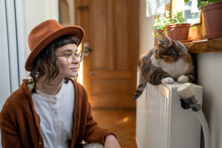 Interested woman cat owner in hat looks to fluffy pet lying on radiator under sunny window at home.の写真素材
