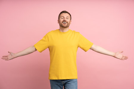Portrait of happy young man spreading arms wide wanting to hug you with closed eyes isolated on pink