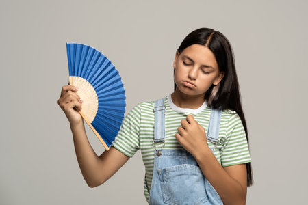 Sweaty exhausted teenage girl using paper fan suffer from heat. Overheating, stuffiness, hot weatherの写真素材