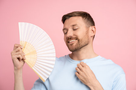 Carefree man using paper fan at summer hot weather enjoying fresh air isolated on studio pinkの写真素材