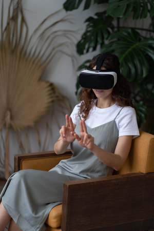 Curious woman in VR helmet touching AR interface with fingers, sitting in urban jungle interiorの写真素材