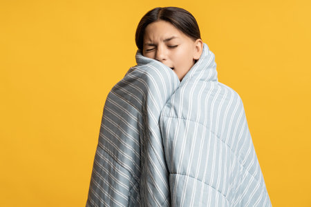 Sleepy teenage girl wrapped in blanket yawns with closed eyes waking up isolated yellow background
