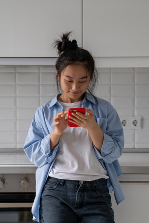 Happy Asian woman holding smartphone reading positive uplifting news while spending time at homeの写真素材