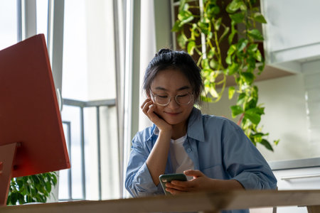 Positive impressed Asian girl student with mobile phone sits at computer desk in home interiorの写真素材