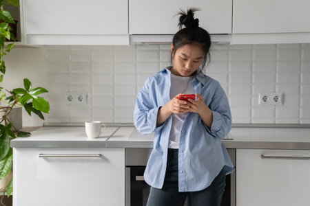 Concentrated emotionless Asian woman typing text on phone standing in kitchen with glass coffeeの写真素材