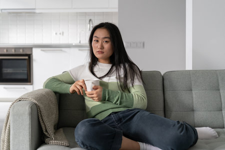 Young relaxed Asian woman looking at camera while sitting on sofa with cup of tea or coffeeの写真素材