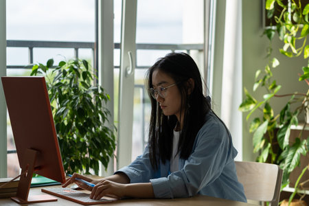 Concentrated Asian woman in glasses internet marketer sit at table work computer in home officeの写真素材