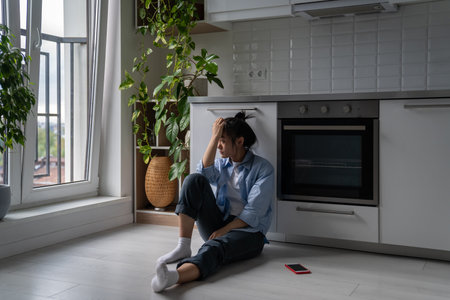 Depressed devastated Asian woman sits near smartphone on home kitchen floor troubled with lonelinessの写真素材