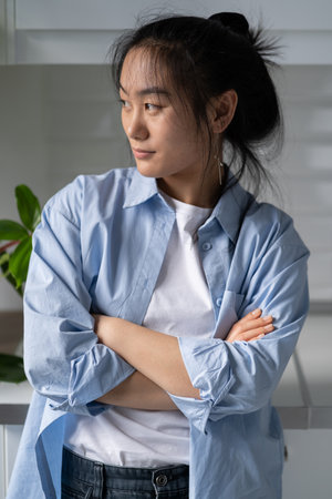 Pensive young Asian girl standing at home with arms crossed looking away thinking of futureの写真素材