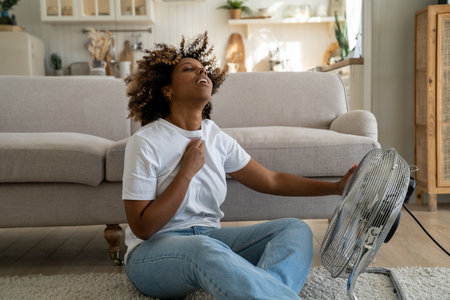 Young African American woman cooling down by ventilator at home, feeling unwell during heatwaveの写真素材
