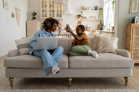 Happy African American family mother and son enjoy weekend at home rest on sofa with gadgetsの写真素材