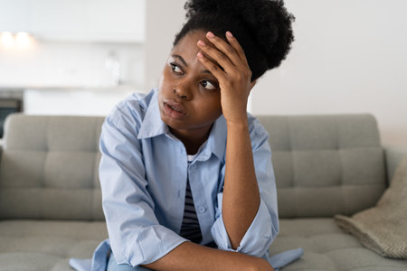 Flustered African American woman is very nervous sighing and touching face with hands sits on sofaの写真素材