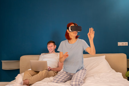 Interested woman in glasses virtual reality sits on bed with husband manages vr goggles from laptop