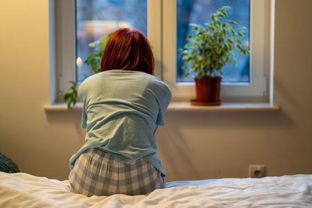 Woman sitting in bed at hone looking at window in evening back to camera.の写真素材