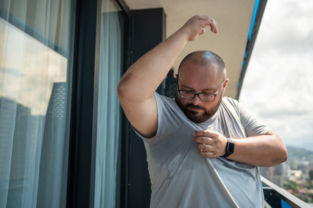 Overweight man suffering from heat, over sweating in summer standing on balcony in summer.の写真素材