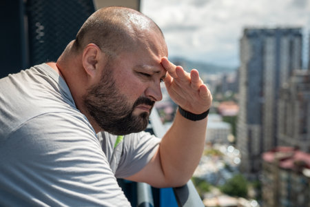 Man covers with hand from sun look into distance from balcony. Fat bald bearded guy hiding with palmの写真素材