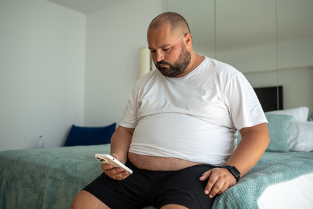 Fat bearded bald guy with smartphone chatting, dials number, types message, watches news, weatherの写真素材
